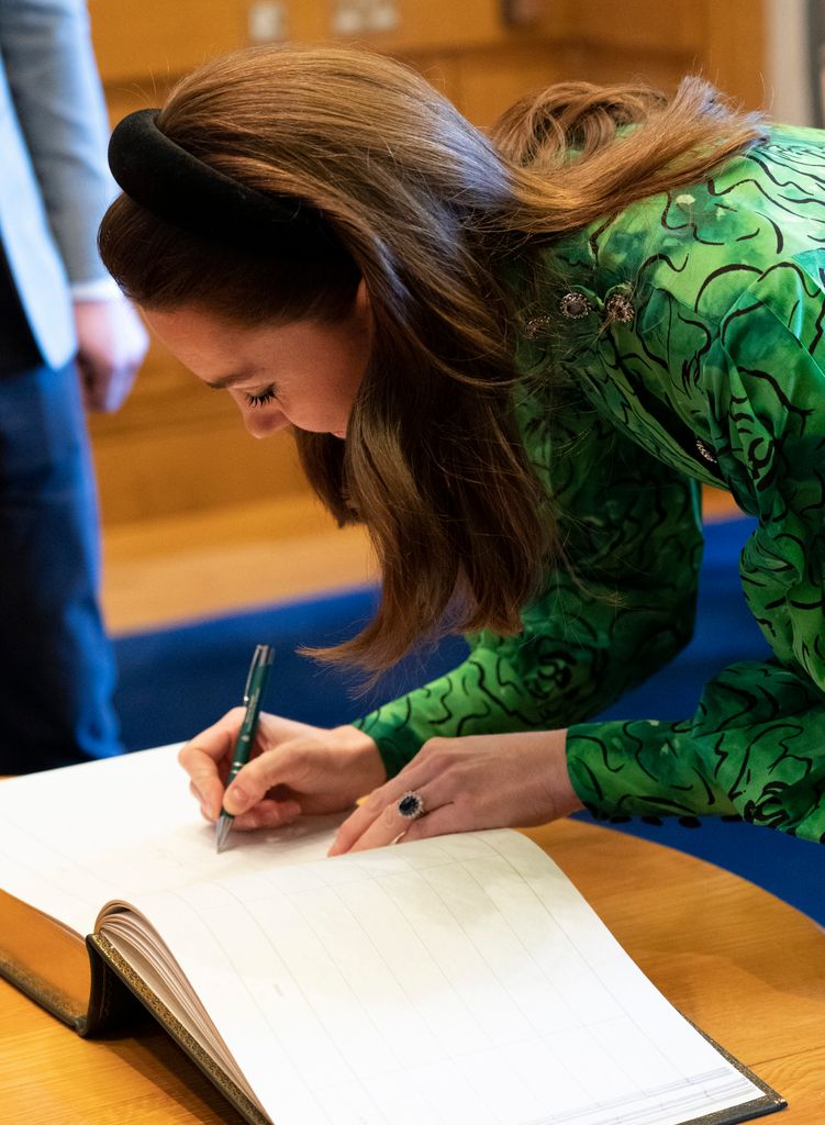 Princess Kate leaning down in a gree dress and headband to sign a big book