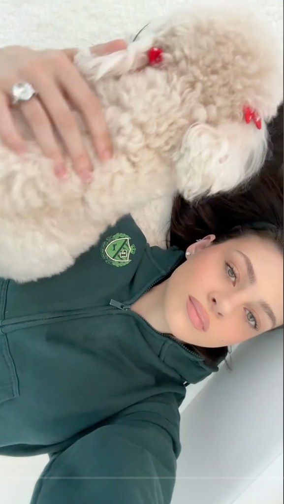 Nicola lies on her back wearing a green hoodie, with her dog resting on her chest. The dog sports adorable red bows, adding a playful vibe.