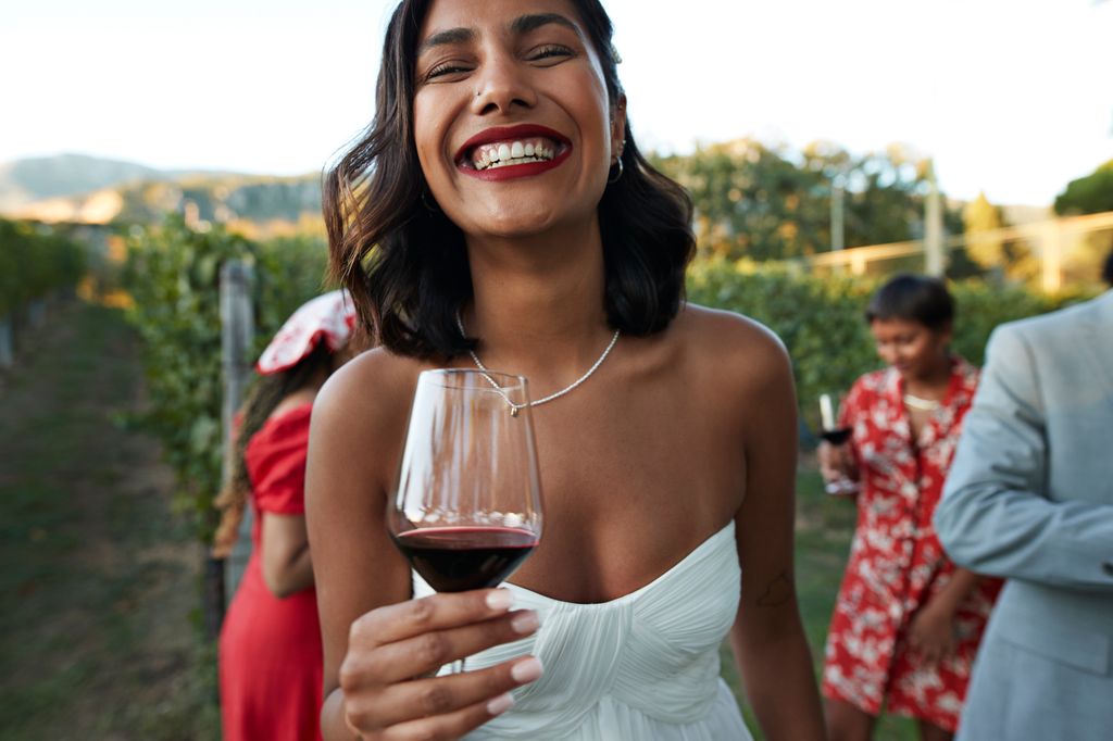 Portrait of happy young bride in off shoulder wedding dress holding wineglass at vineyard