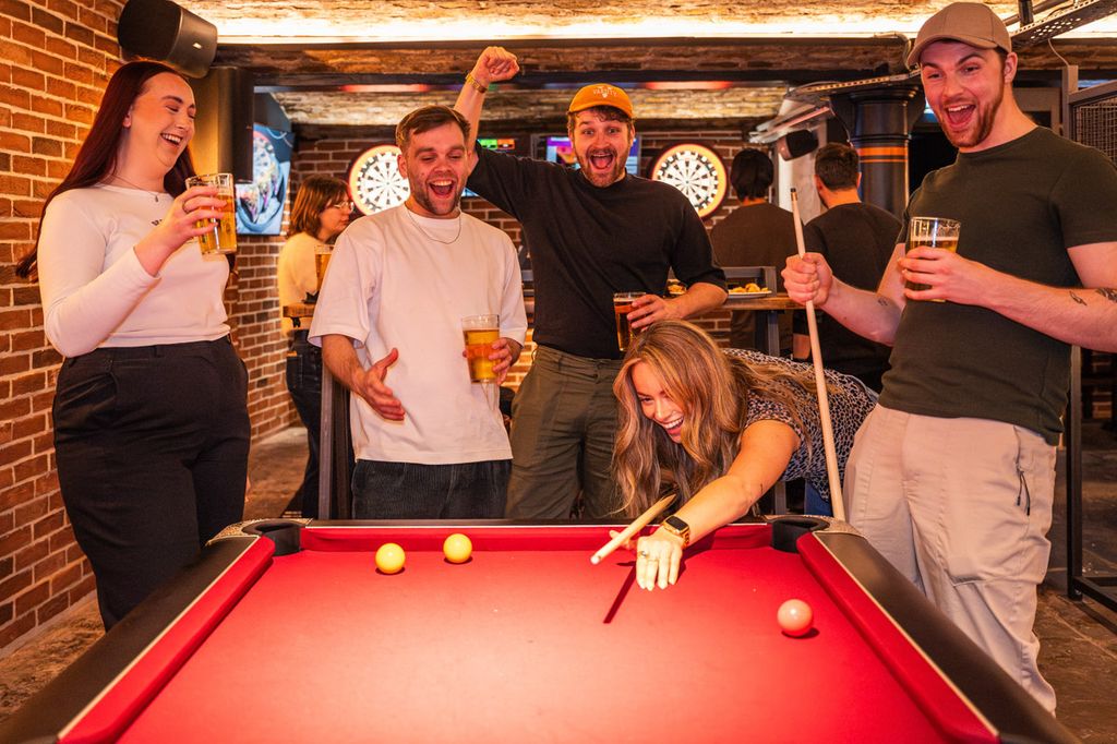 A group of five friends enjoying a lively game of pool in a bar with exposed brick walls and dartboards in the background