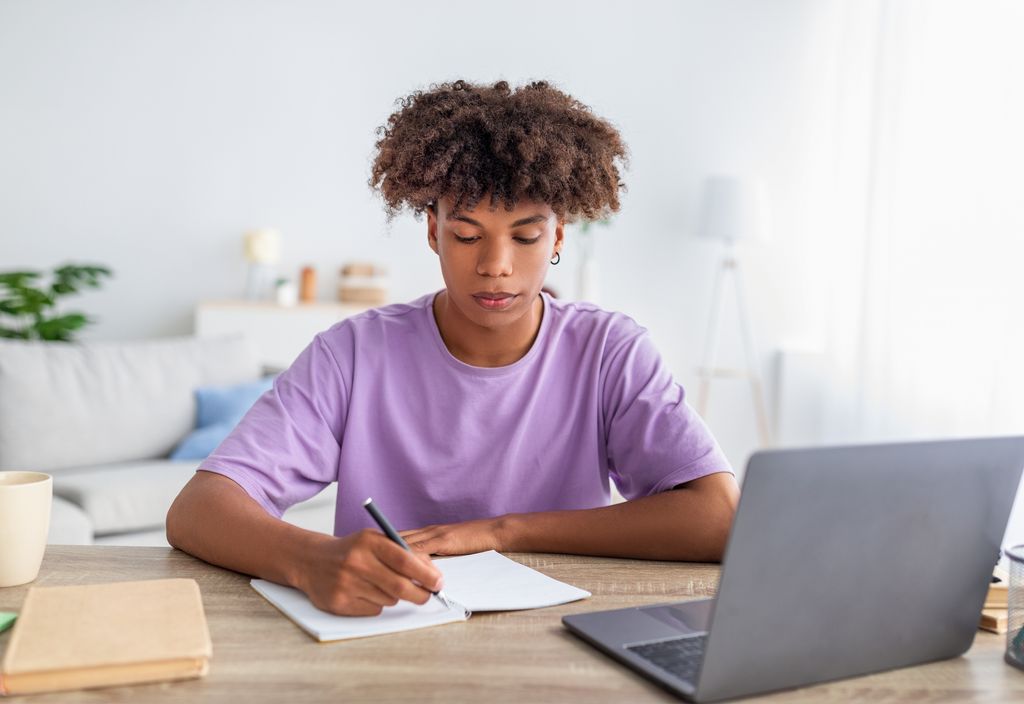 Serious teen studying using laptop and writing in notebook 