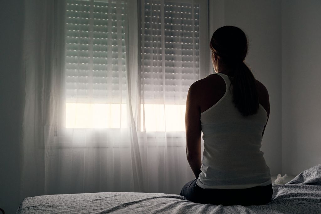 Rear view of an unrecognizable woman sitting on her bed looking out the window.