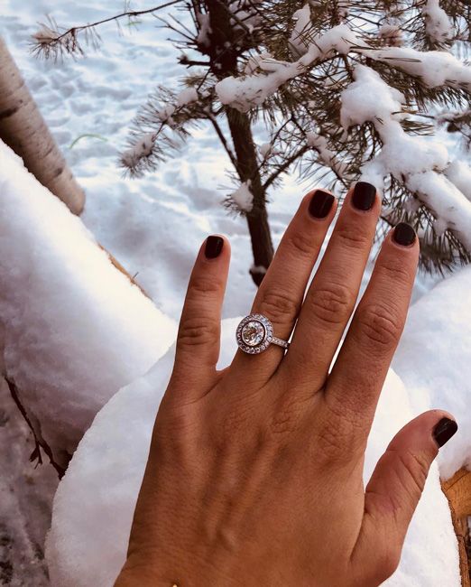 a photograph of a womans left hand wearing a large antique style platinum and white diamond engagement ring featuring one round cut stone surrounded by smaller diamonds and her nails are short with dark polish which contrasts with the pure white snow in the background
