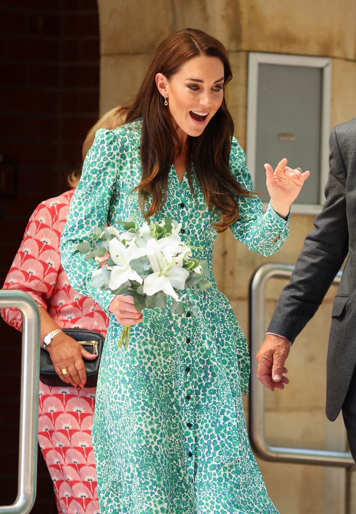 Princess Kate beams with delight, holding a bouquet of white lilies while wearing a green and blue abstract leopard-print maxi dress with puffed shoulders and button detailing. She exudes charm and grace at Riversley Park Children’s Centre.