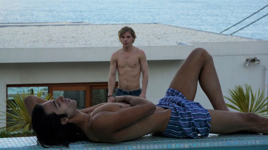 A shirtless man in jeans walkign towards a shirtless man lounging by a swimming pool in trunks