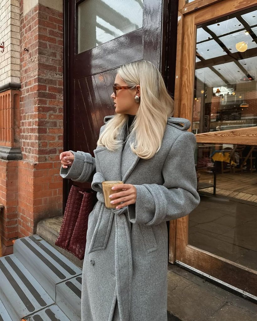 Molly-Mae leans casually outside a rustic doorway, wrapped in a grey wool coat with wide lapels. Her blonde hair, oversized earrings, and sunglasses complement her polished-yet-cozy winter vibe.