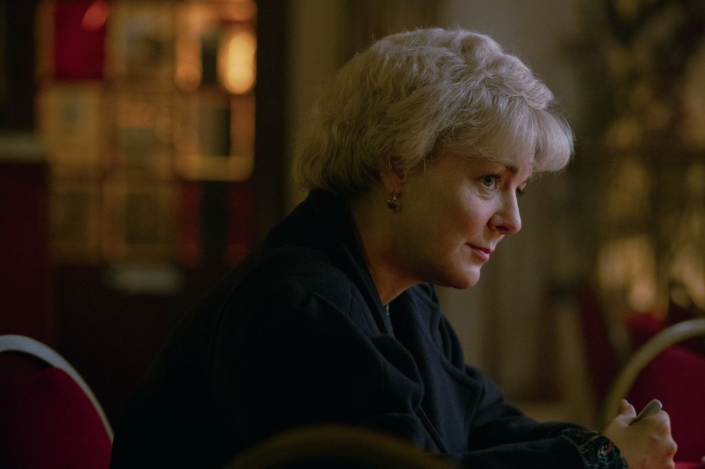 A blonde woman sits with her hands clasped