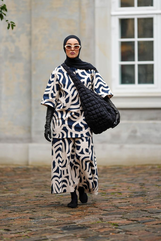 Imane Asry wears a black scarf as a headband, white sunglasses, a navy blue and white latte print pattern oversized t-shirt, matching navy blue and white latte print pattern oversized flowing pants, a black fabric quilted oversized shoulder bag, black shiny leather high gloves, black socks, black shoes , outside Marimekko, during the Copenhagen Fashion Week Spring/Summer 2024 on August 09, 2023 in Copenhagen, Denmark. (Photo by Edward Berthelot/Getty Images)