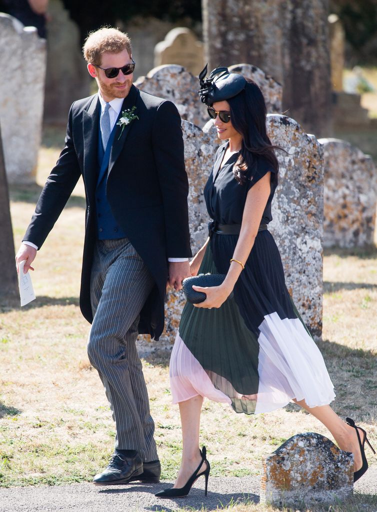 Prince Harry and Meghan Markle walk through a church yard, Prince Harry is wearing a formal suit and Meghan Markle is wearing a colour blocked midi dress and black high heels 