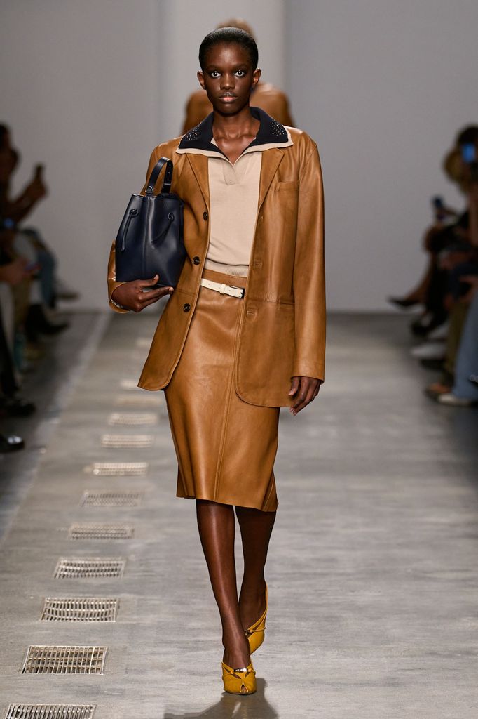 A model wears a tan leather pencil skirt and matching blazer, with a preppy polo top and carrying a navy bag
