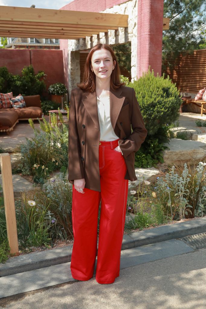 Charlotte Ritchie wears red wide-leg trousers, white blouse and brown blazer stands in garden area at Chelsea Flower Show 2025.