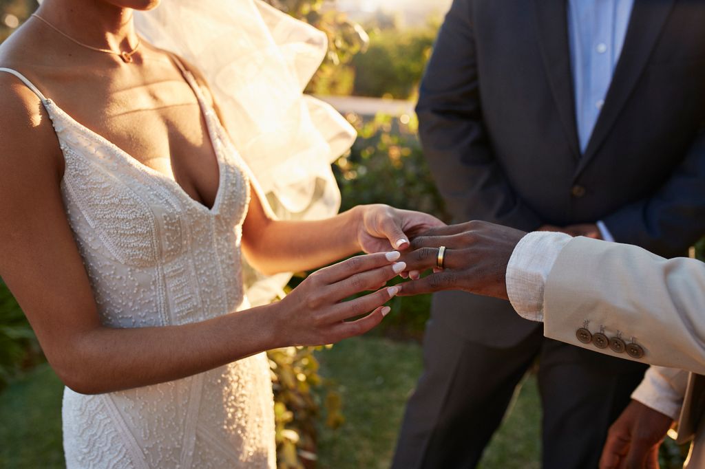 Midsection of young bride exchanging wedding ring with groom while holding hands during ceremony at sunset