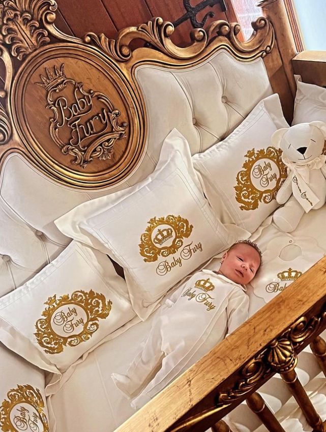 Um bebê em um berço com cinco travesseiros e um ursinho de pelúcia