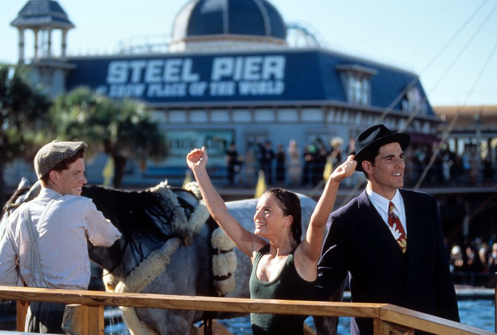 Dylan Kussman porte une casquette plate et une chemise blanche tout en tenant un cheval, regardant Gabrielle Anwar applaudir avec ses bras levés haut. Michael Schoeffling détourne le regard, vêtu d'un costume noir et d'une cravate rouge, avec un chapeau.