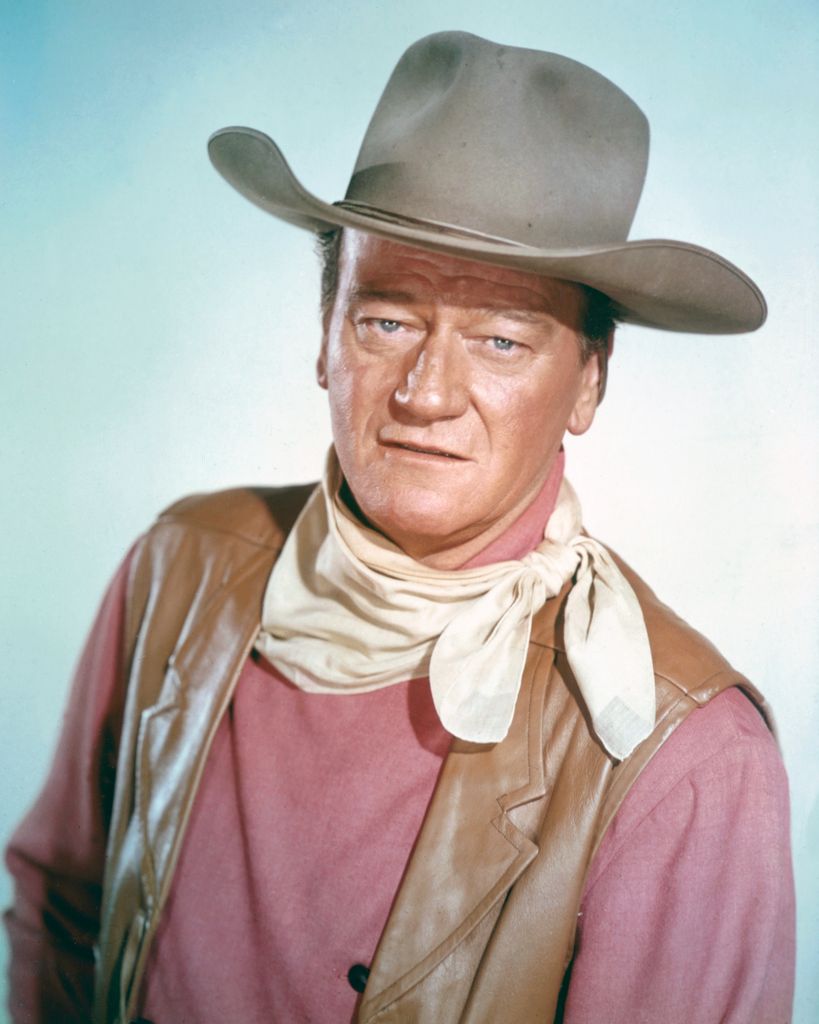 John Wayne (1907 - 1979), US actor wearing a tan leather waistcoat, a pink shirt and a white neckerchief, in a studio portrait, against a light blue background