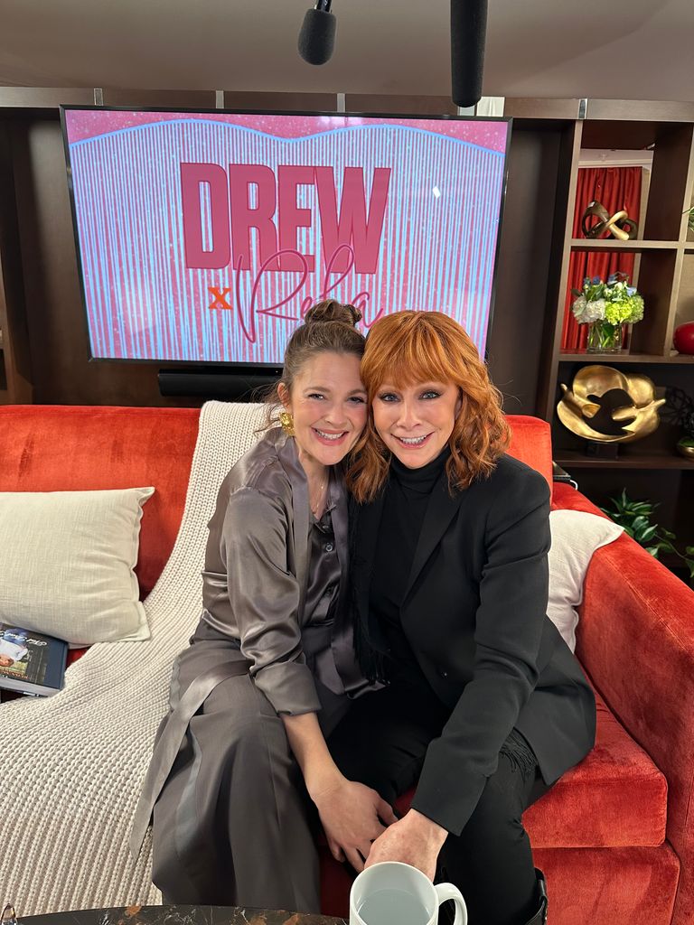 Drew Barrymore and Reba McEntire sit on a red couch and cuddle each other