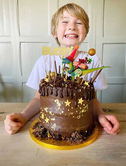 a boy grins at the camera so widely that his top and bottom teeth are showing as a large single tier chocholate cake stands tall in front of him topped with candles and bright sticks of sugary treats