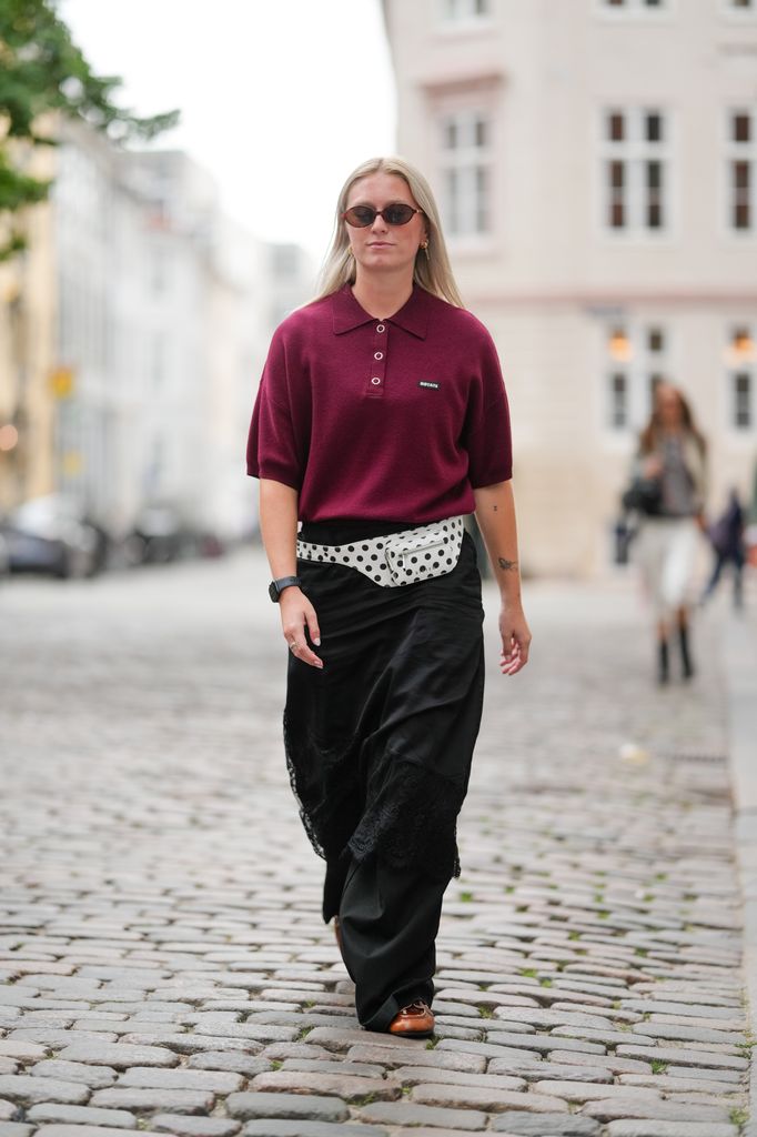  A guest wears straight, center-parted light-blonde hair falling past the shoulders, narrow oval dark-brown sunglasses, and small gold hoop earrings. A deep burgundy knit polo shirt with a spread collar, short sleeves, a three-button placket with white buttons, and a small rectangular black logo patch at the left chest is styled with a white belt bag in a black polka-dot print worn at the waist. A black maxi skirt with a soft drape and tonal lace inset panels toward the hem is paired with brown leather lace-up shoes. A black smartwatch on the left wrist and a ring on the right hand are visible, walking, outside Nicklas Skovgaard, during Copenhagen Fashion Week,