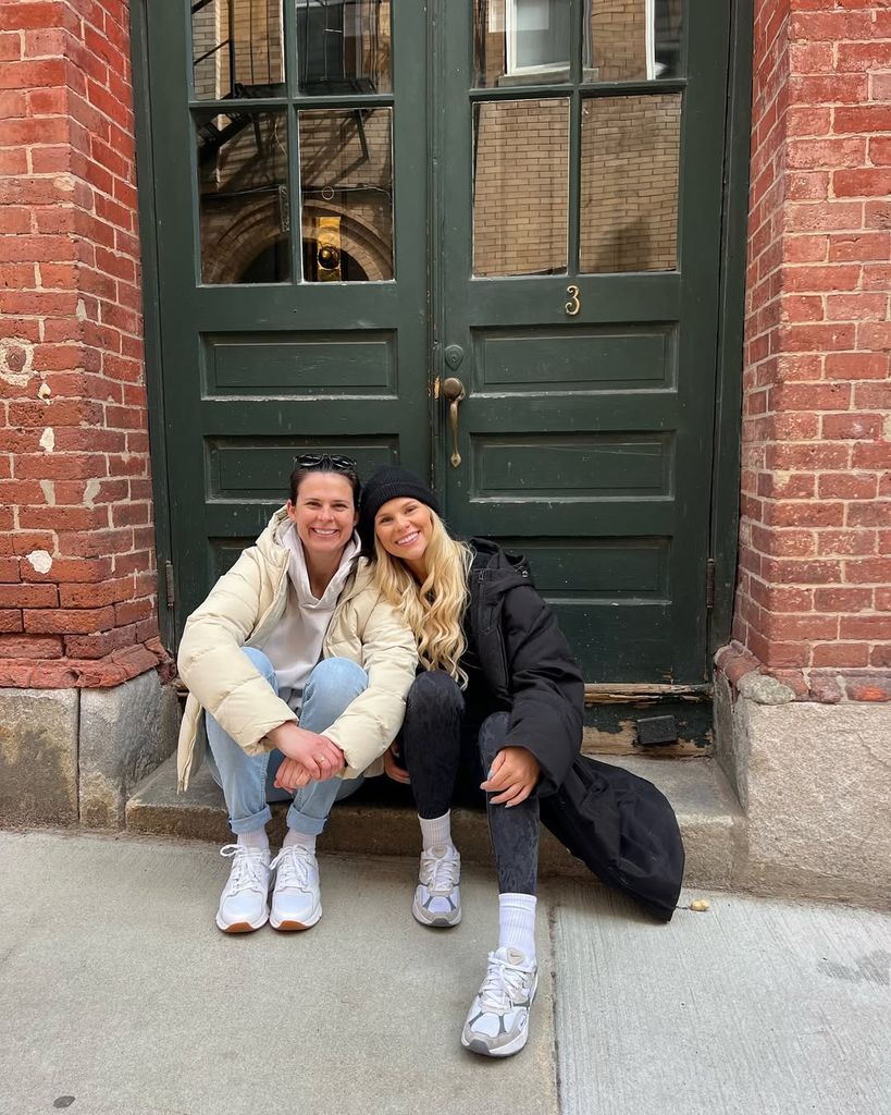 Brittany Bowe et Hilary Knight assises l'une à côté de l'autre sur le pas d'une porte, souriantes et portant des manteaux surdimensionnés.