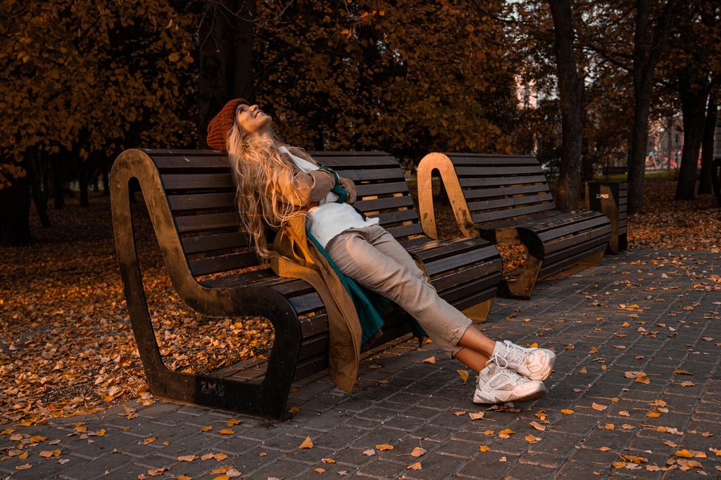 Side view of cheerful young blond female in casual clothes looking up while resting on wooden bench in park in autumn day