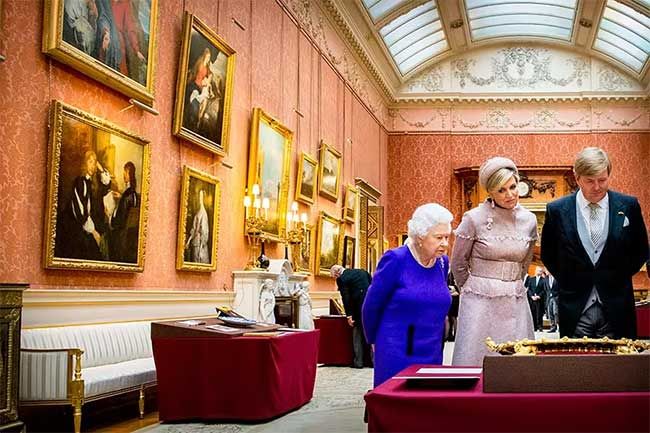 the queen wears a royal blue dress as she stands with a man and a woman observing gold framed oil paintings in a room with a skylight and walls filled with frames from floor to ceiling