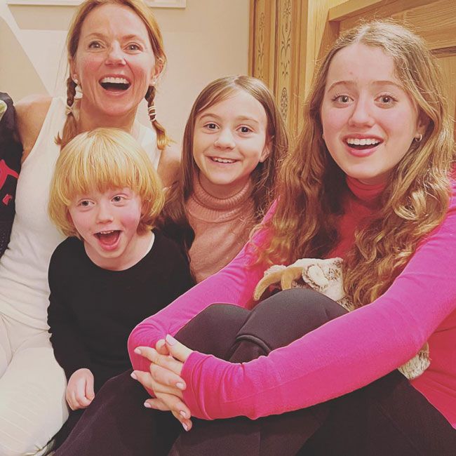 geri smiles at the camera with her young boy and girl in front of her looking happy as a teenage bluebell who is closest to the camera wears a bright pink top and beams with her blue eyes open wide