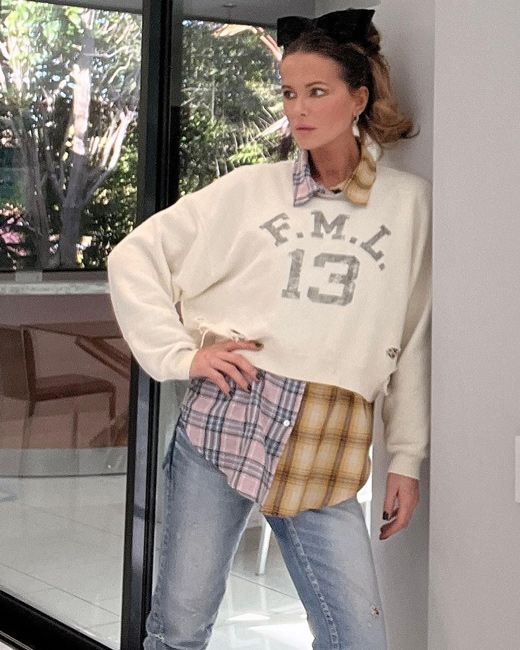 kate stands leaning on a wall with one hand on her hip and wearing a stylish cropped cream sweater with f m l 1 3 written on it