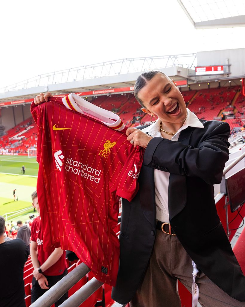 Millie Bobby Brown holding up a Liverpool football shirt at Anfield at Liverpool FC's Premier League-winning game