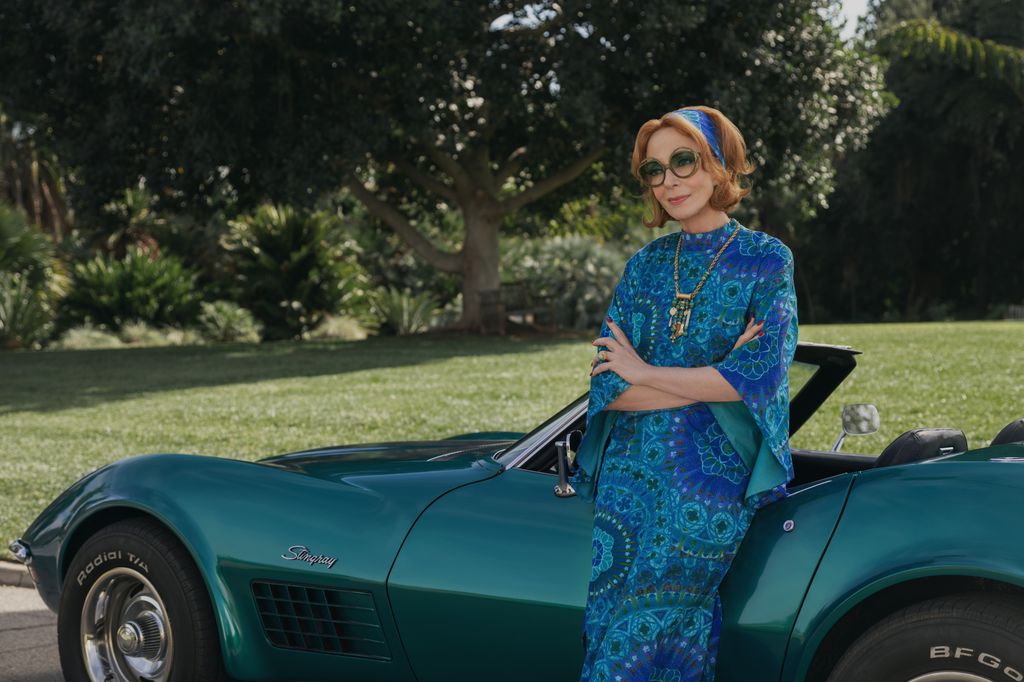 A woman in a blue dress and matching headband leans against a sports car