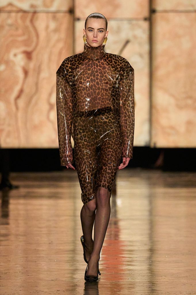 A model walks down the runway at the Saint Laurent show wearing a top and skirt in vinyl leopard print, teamed with gold earrings and sheer tights