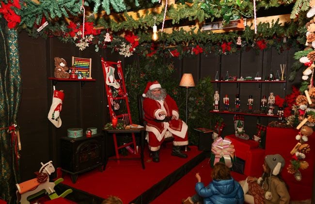 santa sits on a stage inside a grotto pointing at children in the audience