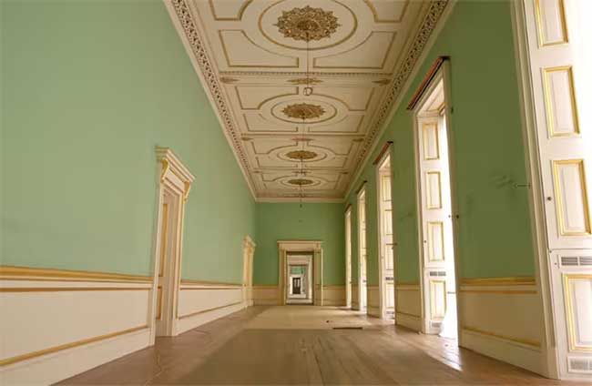 a wide corridor with wooden floors and mint green wallpaper with cream panelling along the lower half and gold detailing running along the edge of each doorway and window positioned every few metres along the way