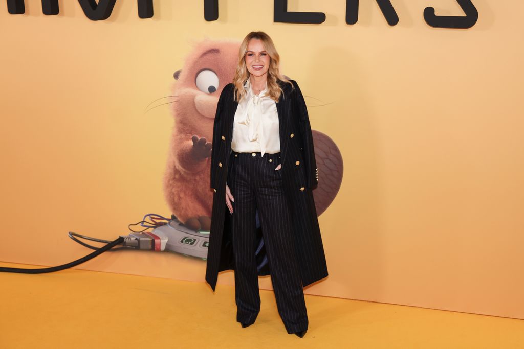 Amanda Holden sourit en portant une chemise blanche Holland Cooper, un pantalon noir et un long manteau noir