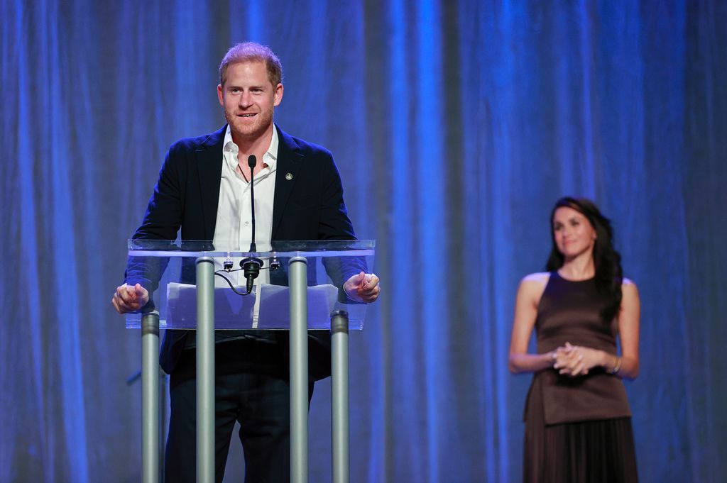 Prince Harry speaking at a podium with Meghan Markle behind them