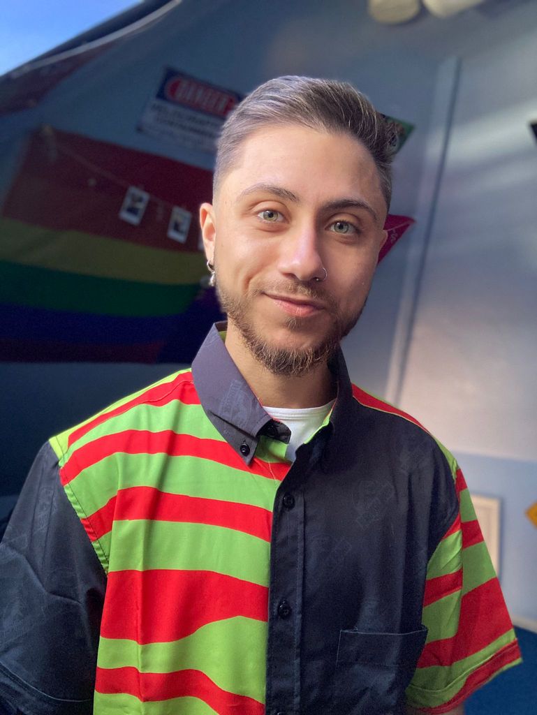A man with facial hair  in a jumper where one half has green and red stripes and the other is a solid blue. In the background is a Pride flag hanging on a wall