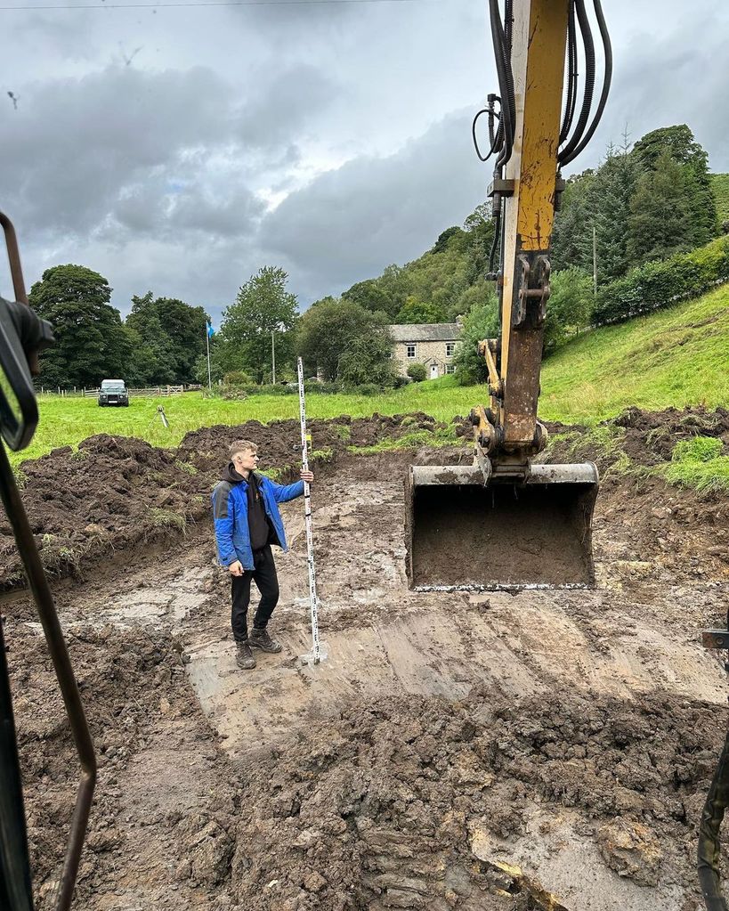 A photo of a young man standing in a muddy hole looking at a large digger