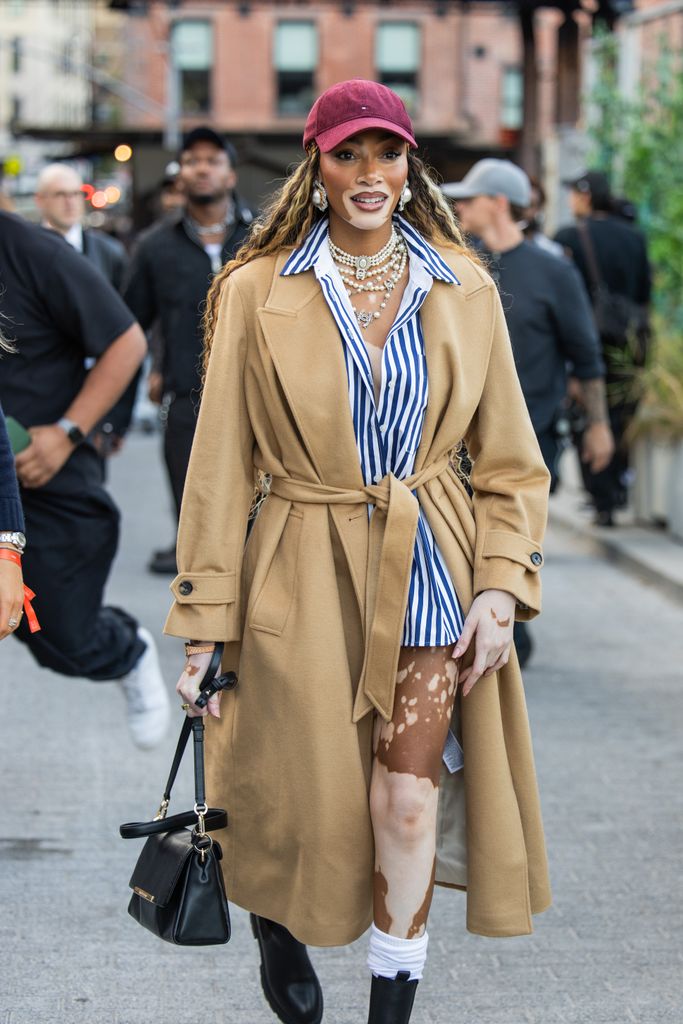 Winnie Harlow wears belted beige coat, striped button shirt, cap outside Tommy Hilfiger New York Fashion Week on September 08, 2024 in New York City. 