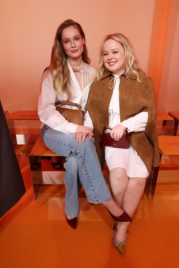 Hannah Dodd and Nicola Coughlan sitting together at the Gucci Women's Spring Summer 2025 Fashion Show. Both look chic—Hannah in her casual jeans and white shirt, and Nicola in her brown cape and white dress. They both sport pointed heels, Hannah's in burgundy and Nicola's in green. The two are smiling, appearing relaxed and engaged.