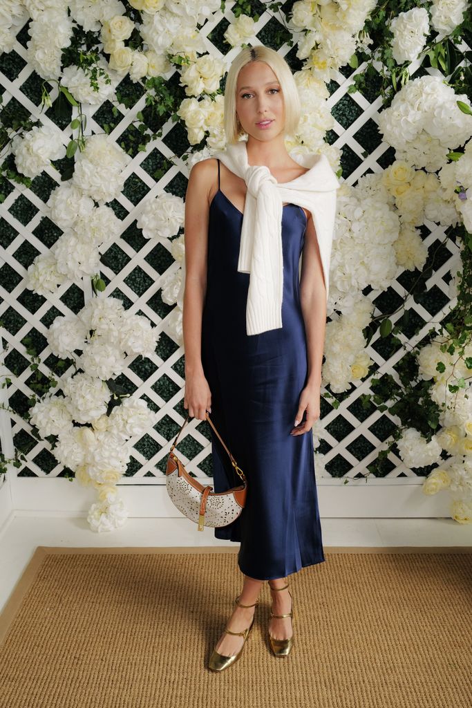 Princess Olympia wears a navy satin slip dress with a cream sweater draped over her shoulders, accessorised with gold shoes and a white shoulder bag