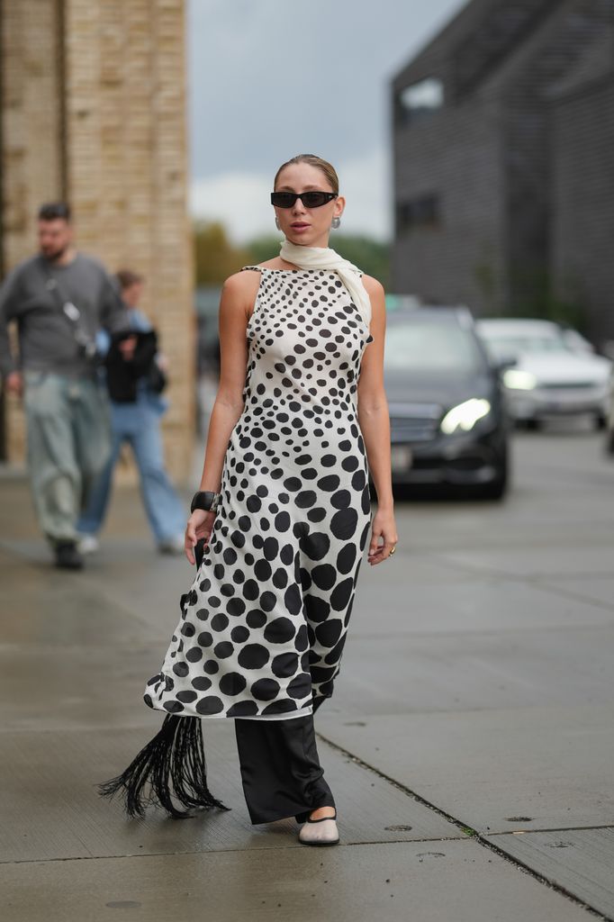  A guest wears a sleeveless dress featuring a black polka dot pattern on a white background, with a halter neck design. A white scarf is draped around the neck. Black rectangular sunglasses are worn. The hair is styled in a sleek, pulled-back bun. A black bangle adorns the wrist. Black wide-leg trousers are visible underneath the dress, which has fringe detailing at the hem. Footwear consists of light-colored flats, outside Gestuz, during Copenhagen Fashion Week