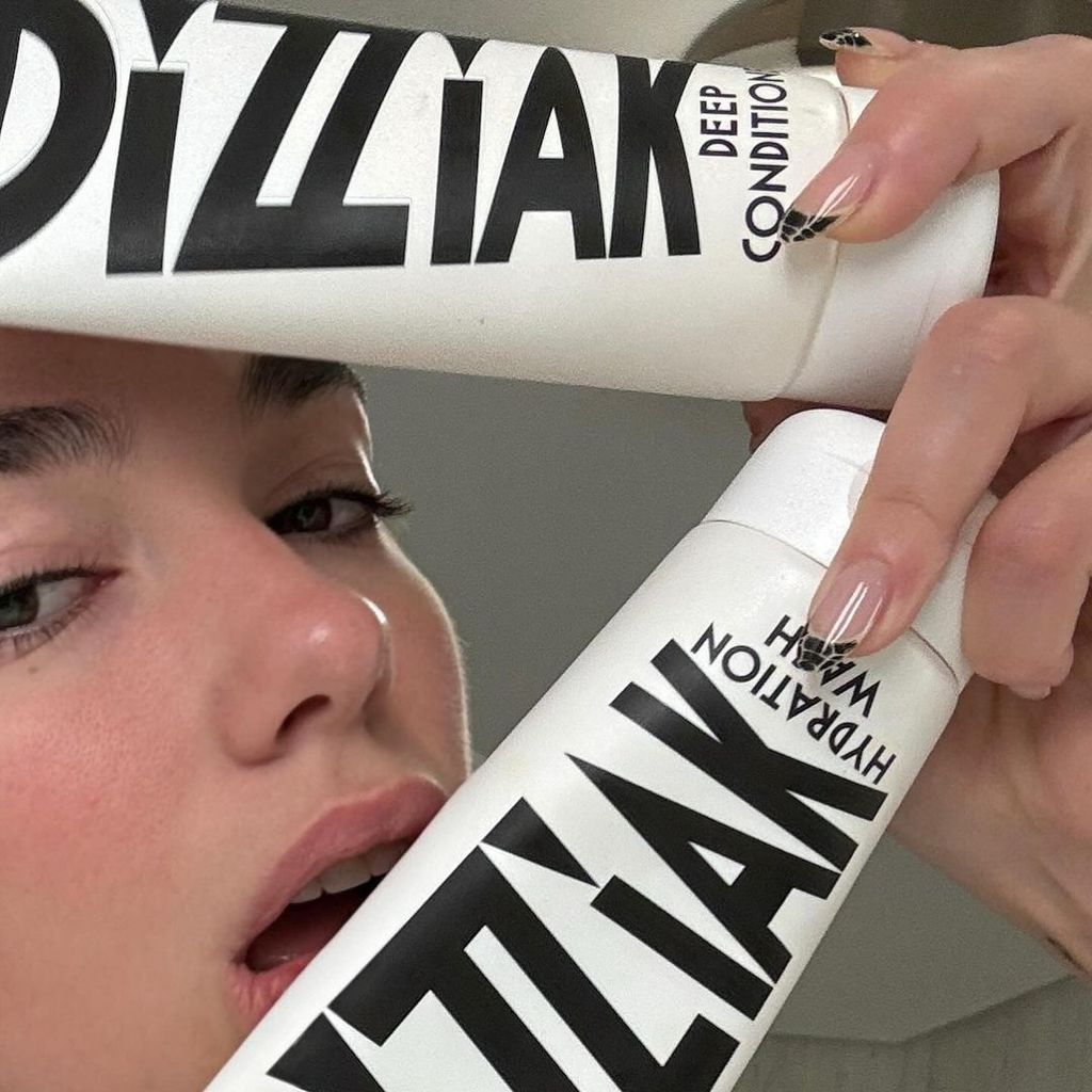 A close-up of Dua Lipa's face as she holds Dizziak's Hydration Wash and Deep Conditioner near her lips, showcasing her natural makeup-free look and stylish, black-and-white nail art.