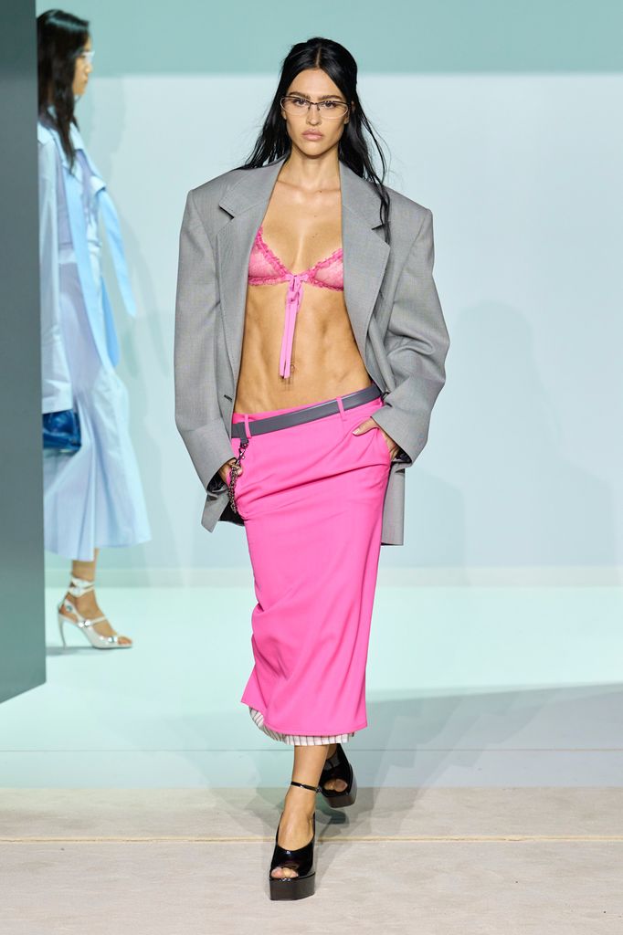 A model wears a pink pencil skirt and bra with an oversized grey blazer and black heels 