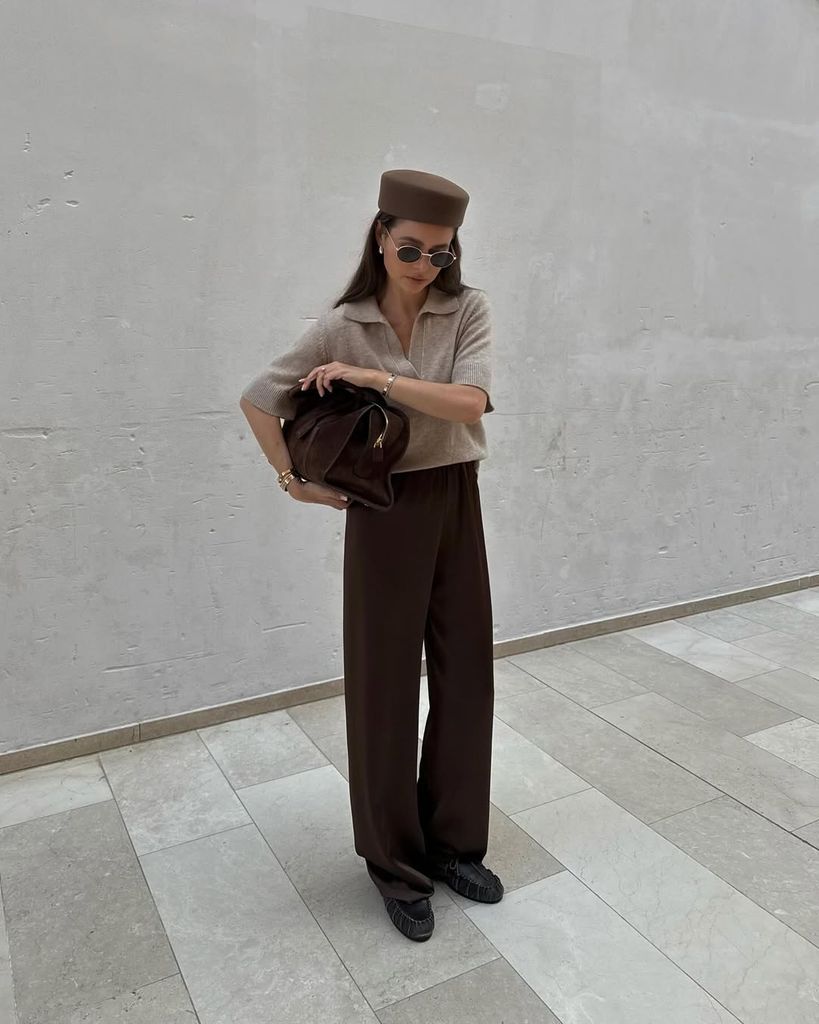 Danish fashion influencer Maria Kragmann poses in a modern vintage-inspired look: taupe knit polo, wide-leg brown trousers, pointed loafers, and a structured pillbox hat. She holds a large brown leather bag.