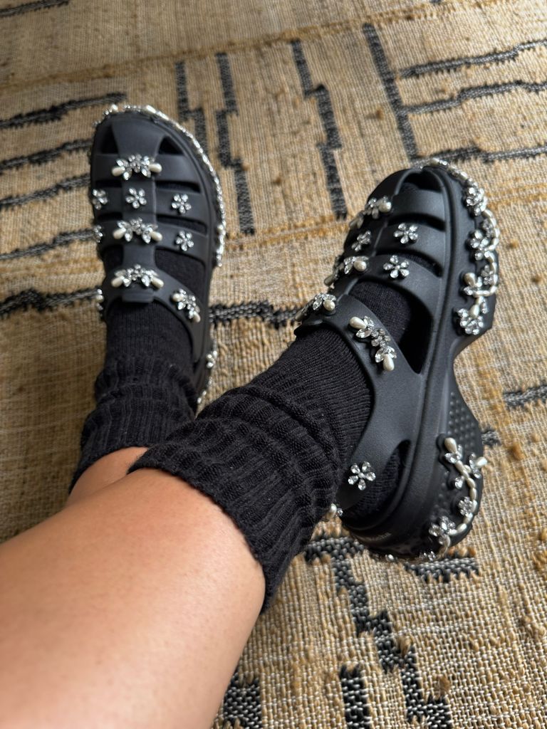 Close-up of Nieve Tierney's feet in black platform sandals adorned with pearl and flower embellishments, styled with black socks, resting on a woven beige and black rug.