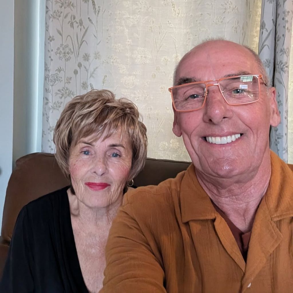 Dave and Shirley Griffiths take a selfie in their living room with floral curtains in the background