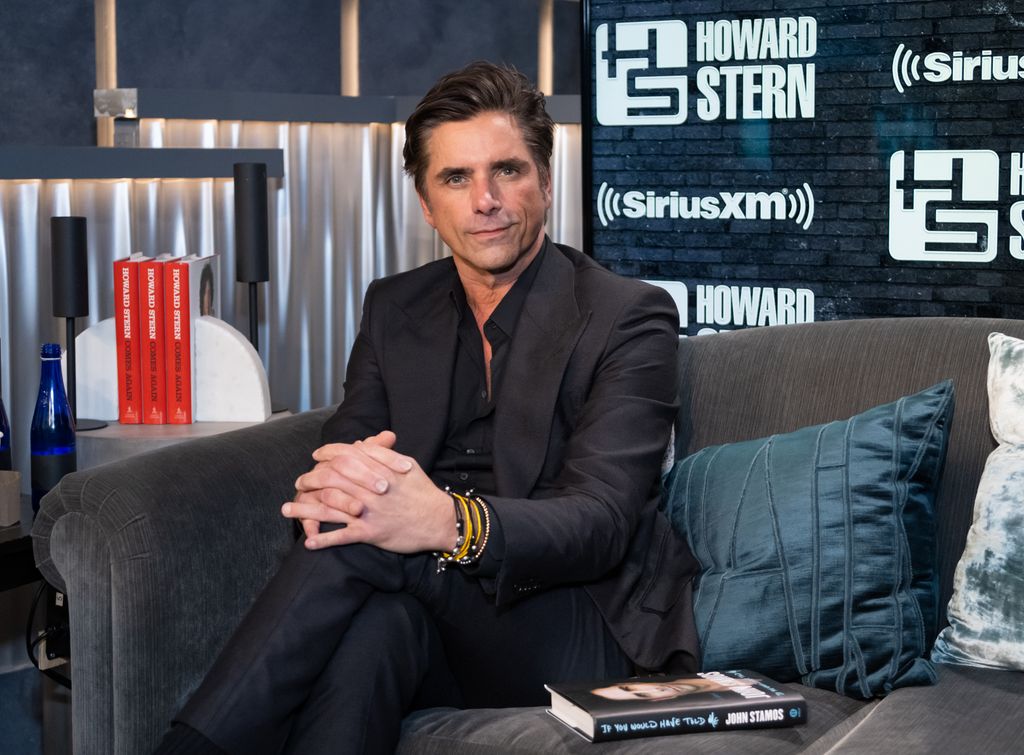 john stamos in dark suit sitting on couch