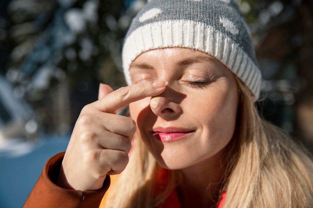 blonde woman wearing knit hat applying sunscreen to nose outside in winter