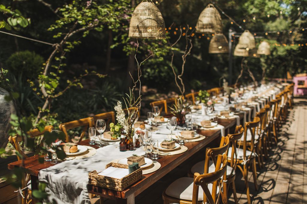 Trendy and beautiful table setting for romantic wedding dinner in rustic style. Food catering outdoors. Wooden furniture, lamps and bulbs.