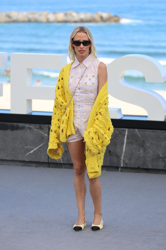 Olivia Baglivi stands on an outdoor platform in front of the ocean. They wear a pastel plaid romper with a yellow chunky knit cardigan and cream-colored flats. Sunglasses complete the beachy look.