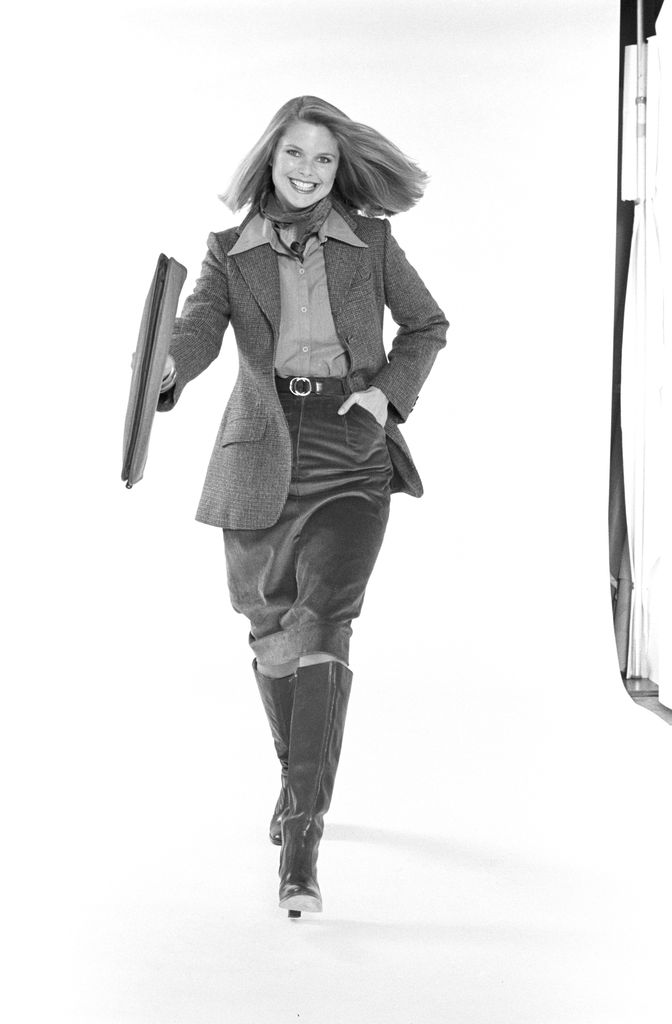 American model (and later actress) Christie Brinkley poses, dressed in business attire, against a white background, 1976. She wears a neckerchief, a tweed jacket over a button-down blouse, a corduroy skirt, and boots.
